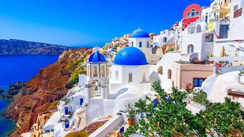 (TAP) - Santorini - Thiên đường du lịch giữa biển Aegean là hòn đảo nổi tiếng nhất trong quần đảo Cyclades thuộc Hy Lạp. Nơi đây mê hoặc du khách bằng khung cảnh như tranh vẽ: những ngôi nhà trắng xanh nằm cheo leo trên vách núi, biển xanh biếc vỗ về bờ cát đen độc đáo.  Theo Cổng thông tin Tổ chức Du lịch Santorini (santorini.gr), Santorini được kiến tạo từ vụ phun trào núi lửa Minoan khoảng 3.600 năm trước. Hòn đảo nằm ở phía Đông Nam thủ đô Athens khoảng 200km, có diện tích gần 76 km² và là nơi sinh sống của hơn 15.000 cư dân. Hành trình khám phá Santorini sẽ trọn vẹn nhất trong khoảng tháng 4 - tháng 10, khi hòn đảo chìm trong tiết trời khô ráo, biển lặng sóng và nắng vàng dịu dàng.  Đến thăm Santorini sẽ thật thiếu sót nếu bỏ qua làng Oia, nơi các ngôi nhà trắng muốt, mái vòm xanh lam xếp tầng trên vách núi cheo leo, tạo nên khung cảnh hùng vĩ, lãng mạn đặc trưng. Ngoài ra, du khách có dịp chiêm ngưỡng khung cảnh hoàng hôn rực đỏ trên nền trời, dạo bước qua các gian hàng thủ công tinh xảo, thưởng thức ẩm thực Địa Trung Hải ngay tại nhà hàng nép mình bên sườn núi. Sự yên bình cùng kiến trúc Cycladic đặc trưng khiến Oia trở thành điểm check-in hấp dẫn. Nếu Oia mang cảm giác tĩnh lặng, trầm mặc thì Thira (hay Fira) - trái tim của Santorini lại cuốn hút bởi nhịp sống sôi động với vô số quán cà phê, nhà hàng, bảo tàng và cửa hiệu lưu niệm. Từ đây, du khách có thể di chuyển bằng cáp treo hoặc đi bộ xuống bến cảng cổ Ammoudi để khám phá vịnh nhỏ xinh đẹp, bãi biển cát đen huyền bí; đồng thời tìm hiểu văn hóa, lịch sử địa phương qua nhà thờ mái vòm, phòng trưng bày nghệ thuật đầy cảm hứng.